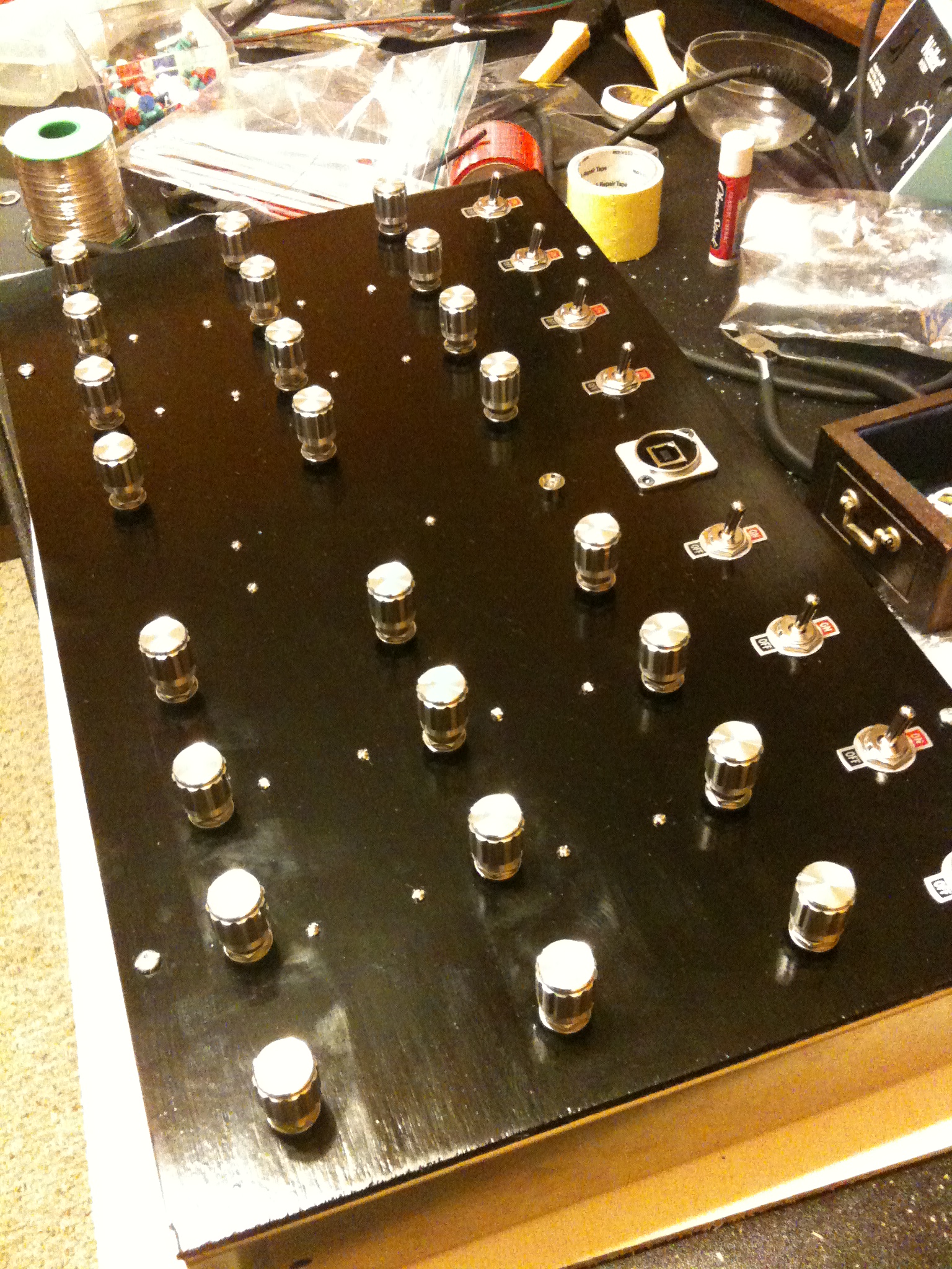 A large black panel densely populated with silver knobs and toggle switches, under construction on a cluttered bench