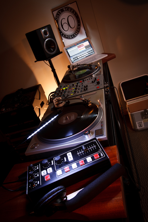 A dim studio with turntables, a laptop, a Sixty Works poster on the wall and a controller in the foreground