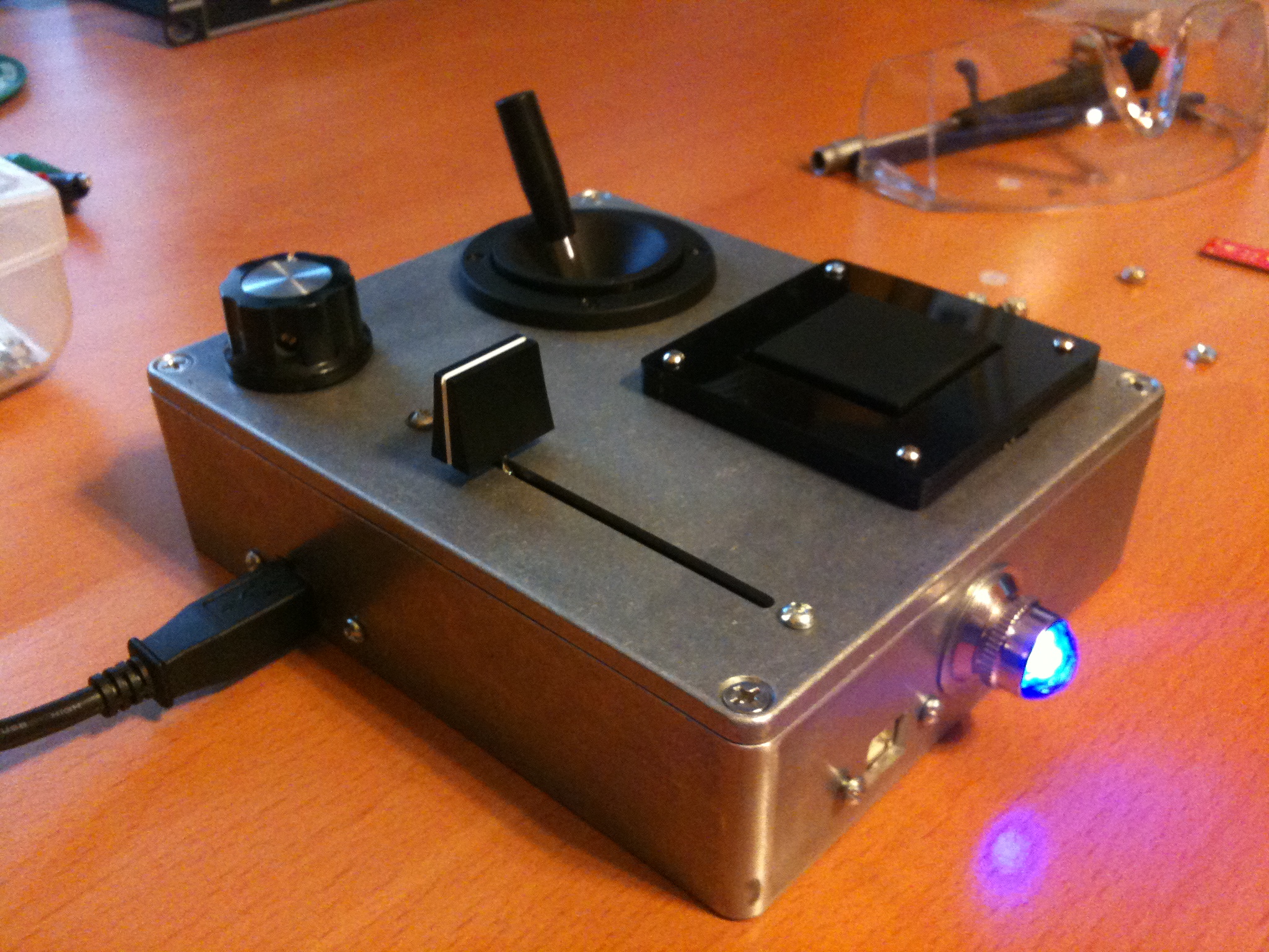 Aluminum enclosure controller with a joystick, fader, knob and trackpad, USB cable attached, on a wood desk