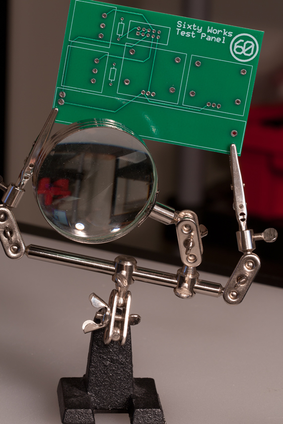A green Sixty Works Test Panel PCB held in a helping-hands clamp behind a magnifying glass